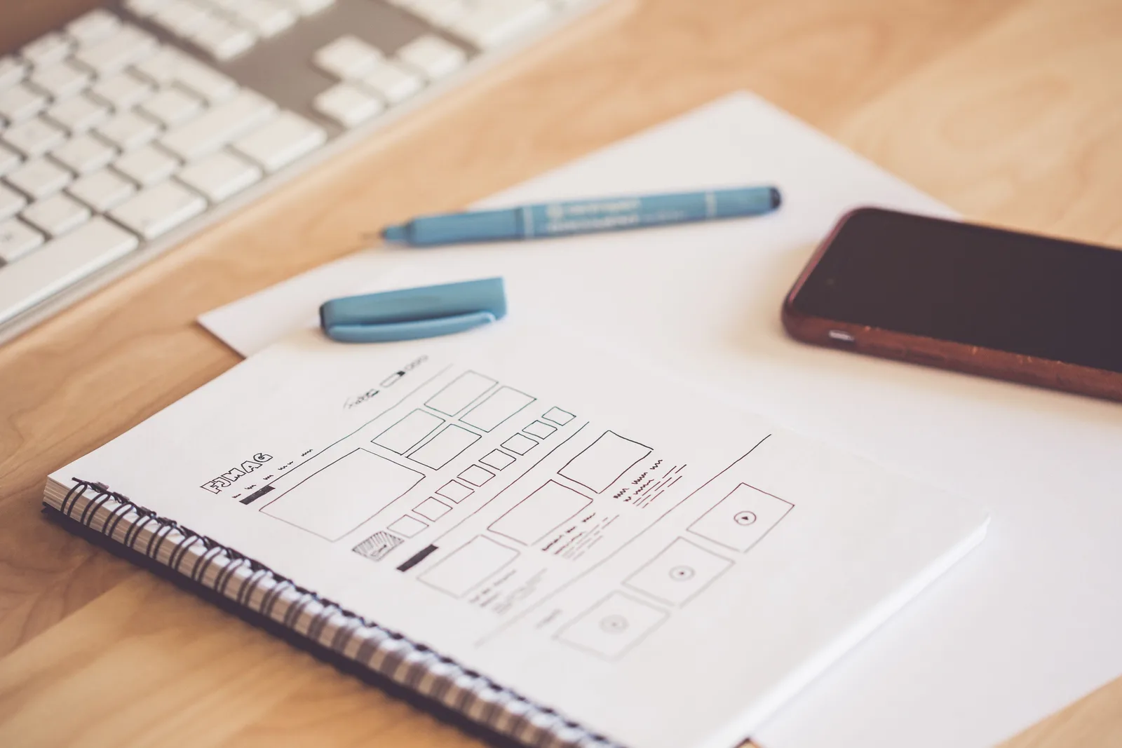 A neat workspace featuring a sketchpad with wireframes, a smartphone, and a keyboard on a wooden desk. Product marketing roles and responsibilities by team