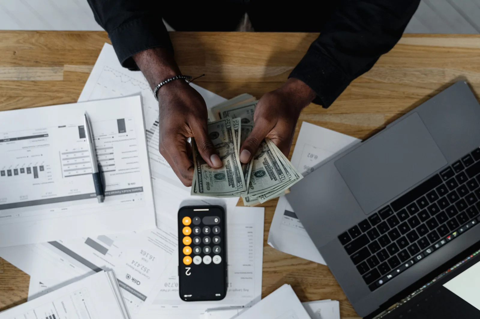 Person counting US dollars, using a calculator and laptop, with financial documents on a wooden desk. Making sense of product marketing salary